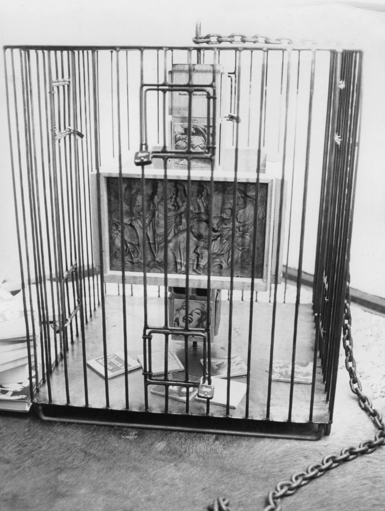 A metal cage with small doors locked by padlocks. The totem-like pole inside contains collaged photography and small wood reliefs.