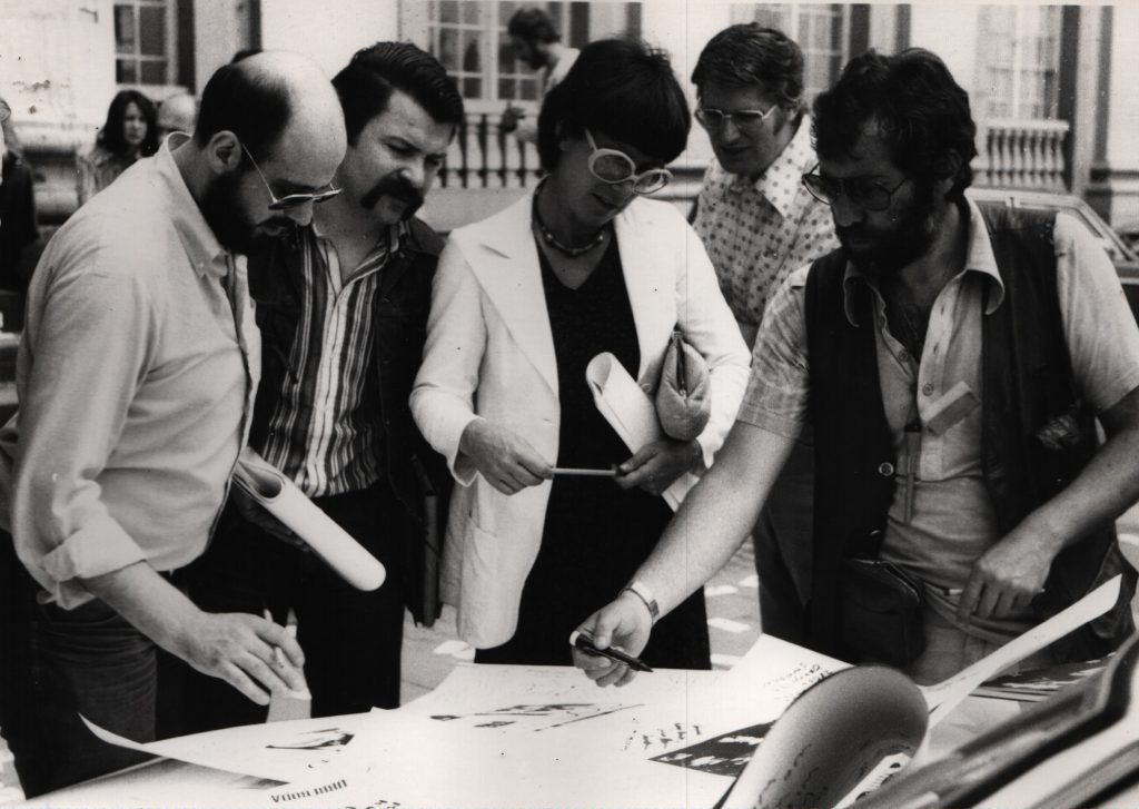 Persons stading in a circle on the street in front of engine hood. 1 man explains a large white sheet lying on the engine hood.
