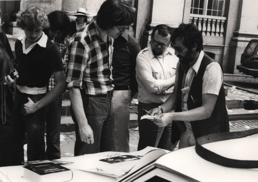 2 man stand in street in front of engine hood. 1 explains something to the other.
