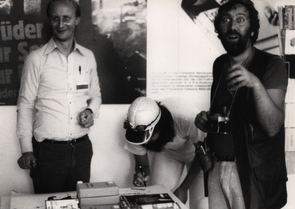 3 mans stand beside table. 1 man writes on large white sheet with collages.