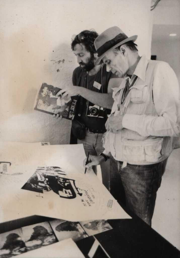2 man stand beside table.. 1 man writes on white sheet with collages lying on the table..