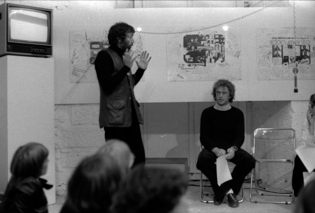 Man standing in a room and talking with other persons sitting on chairs.